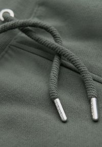 Close-up of two dark green drawstrings with silver metal tips on a fabric surface, likely part of a hoodie or sweatpants.