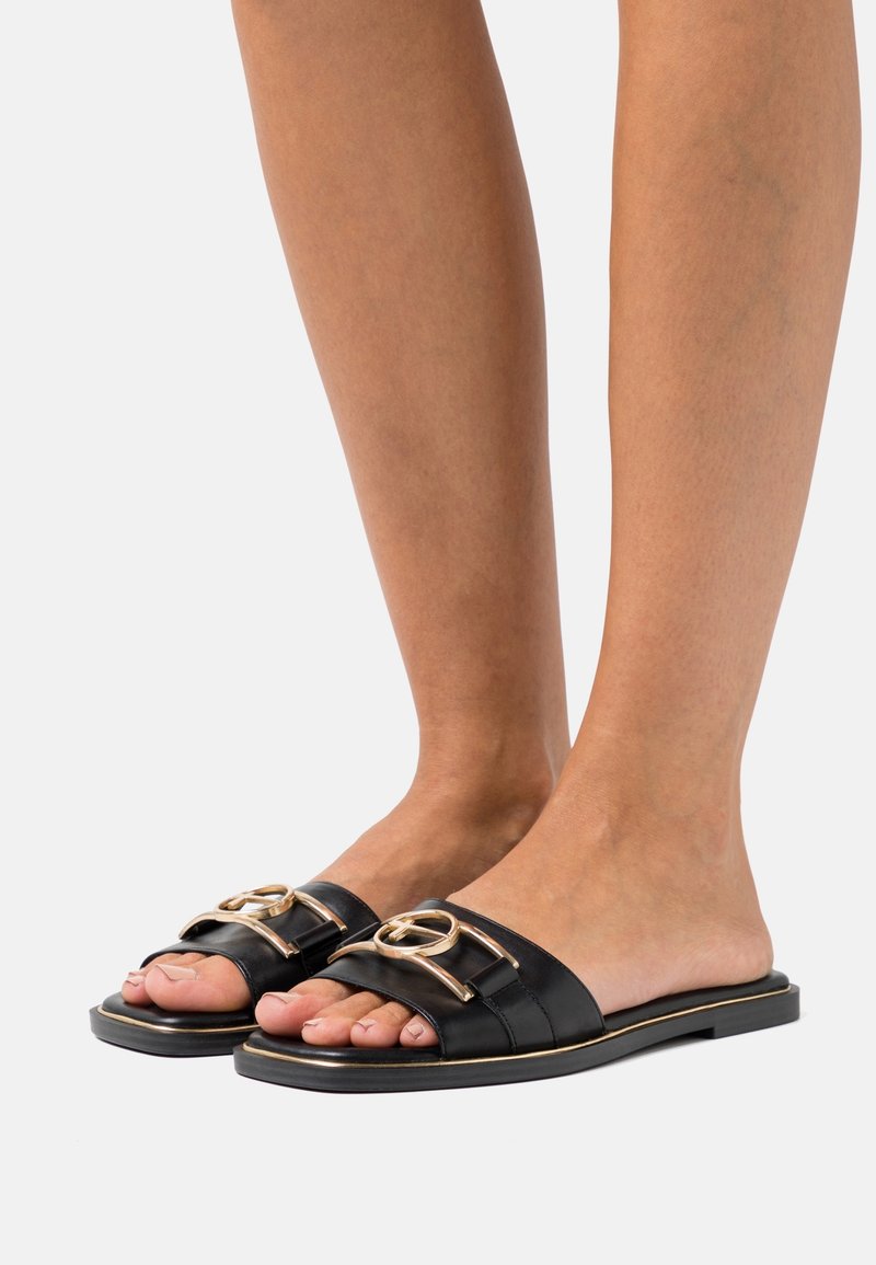 Sandales noires en cuir avec deux brides et accents en métal doré. Semelle plate et style à bout ouvert, montrant les orteils.