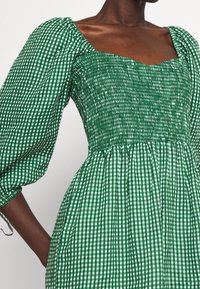 Zelené kostkované šaty s nafouklými rukávy, smockovaným korzetem a čtvercovým výstřihem. Tkanina má kostkovaný vzor a texturovaný povrch.