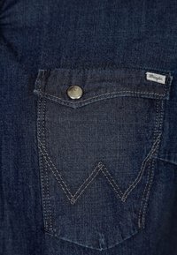 Mörkblå denim med en ficka som har en distinkt zigzag-söm och en silverfärgad knapp, märkt med en liten "Wrangler"-tag.