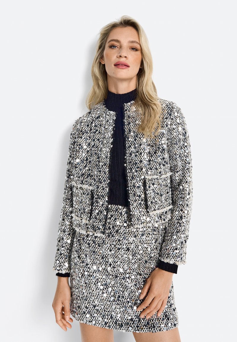 Sequin jacket with a cropped design, a dark knitted collar, front pockets, and a metallic color scheme of silver, black, and white.