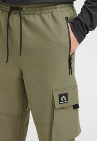 Los pantalones cargo verde olivo cuentan con una cintura con cordón, bolsillos con cremallera, un bolsillo lateral de parche y una etiqueta de logo negra en el muslo.