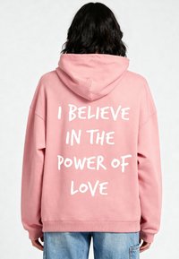 Felpa con cappuccio rosa dal taglio comodo, dotata di una tasca a marsupio frontale e di una scritta bianca sulla schiena che recita "CREDO NEL POTERE DELL'AMORE."