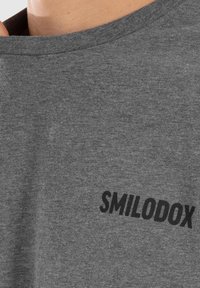 Camiseta gris con cuello redondo y mangas cortas, que presenta un logo negro de "SMILODOX" en el frente, fabricada en un tejido suave y texturizado.