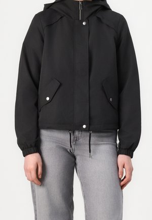 Sort hættetrøjejakke med frontlynlås, lommer med trykknapper, elastiske manchetter og snore, parret med lysegrå jeans.