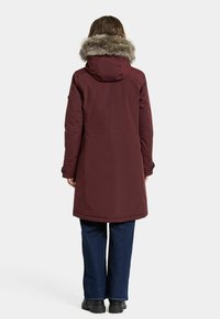 Bordeaux parka med en hætte af kunststofpels. Har en glat tekstur, lang længde og sømdetaljer. Båret over blå vide jeans og sorte støvler.