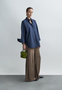 Laivastonsininen liituraitakauluksellinen paita, jossa on leveät hihat, yhdistettynä tekstuuriin ruskeissa leveälahkeisissa housuissa. Vihreä käsilaukku lisää väripilkun.