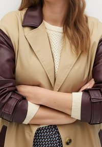 Trench beige con maniche in pelle marrone, dotato di colletto a revers e chiusura con bottoni. Maglione in maglia sotto con motivo a rilievo.