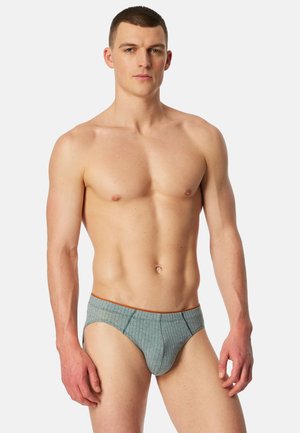 Junger Mann mit kurzen Haaren, der graue karierte Unterhosen mit orangefarbenem Rand trägt und vor einem schlichten hellen Hintergrund steht.