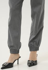 Pantalons de jogging gris avec des poignets élastiques associés à des mules à talons noirs avec des pointes et un détail de boucle décoratif. Texture de tissu lisse.
