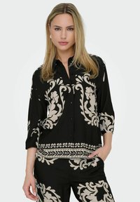 Blouse noire avec des motifs floraux beiges, devant à boutons et manches trois quarts. Tissu doux et lisse avec des détails délicats le long de l'ourlet.