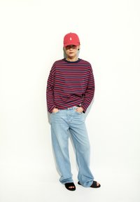 Rot-blau gestreiftes Langarmhemd, hellblaue weite Jeans, schwarze Riemchen-Sandalen und eine rosa Mütze mit weißen Banddetails.
