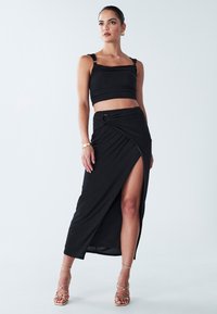 Crni dvodijelni outfit koji se sastoji od uskog crop topa sa širokim trakama i suknje visokog struka. Suknja ima bočni prorez i detalj čvora.