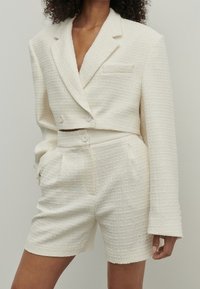 Valkoinen teksturoitu puku, joka sisältää lyhytmallisen, kaksirivisen bleiserin ja korkea vyötärö shortsit, joissa on sivutaskut ja nappikiinnitys.