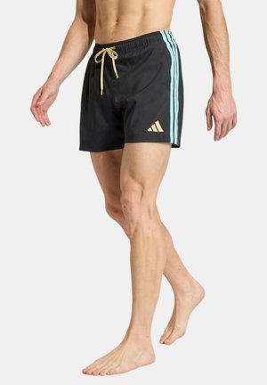 Hombre con pantalones cortos de baño negros con franjas laterales azul claro y cordón beige, de pie descalzo sobre fondo liso.