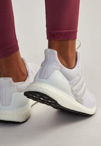 Baskets de sport blanches avec dessus en mesh, trois bandes et semelle en mousse dense. Étiquetées "ultra boost" près du talon. Portées avec des leggings bordeaux.