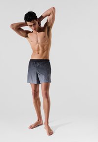 Pilkos maudymosi šortai su juodu elastingu juosmeniu, lygus paviršius ir šoninė grafikų dizaino detalė. Modelis pozuoja basomis kojomis, su rankomis už galvos.