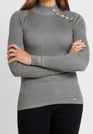 Femme portant un pull gris ajusté à côtes avec un col montant, des boutons décoratifs dorés sur l'épaule et des rayures dorées sur une manche, bras croisés.