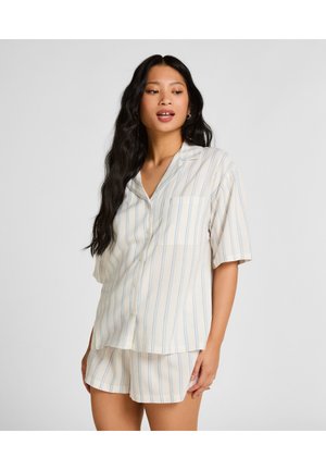 Frau mit langen, welligen Haaren, die ein cremefarbenes, kurzärmliges Pyjama-Set mit dünnen blauen vertikalen Streifen trägt.