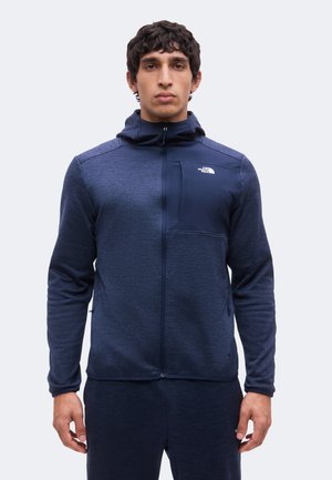 Mann schaut nach vorne und trägt eine dunkelblaue Kapuzenjacke mit Reißverschluss, einer Brusttasche und dem The North Face-Logo auf der linken Seite.