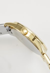 Montre dorée avec un boîtier rond, bracelet en métal argenté, couronne proéminente et surface lisse et polie. Design classique avec un minimum de détails.