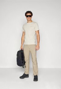 Muž v béžovém tričku, khaki kalhotách, černých teniskách a velkých černých slunečních brýlích, držící černý batoh na jednobarevném pozadí.