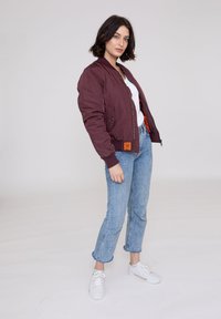 Bordeaux bomberjakke med ribbede manchetter, lynlås fortil og sidelommer; båret over en hvid top, parret med lyseblå jeans og hvide sneakers.