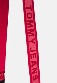 Cinturino rosso con le parole "TOMMY JEANS" intrecciate in un rosa chiaro a contrasto. Il materiale sembra resistente con una texture liscia.