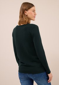 Jeune femme aux cheveux bruns à shoulder longueur, portant un pull à manches longues vert foncé et un jean bleu, de profil devant un fond uni.