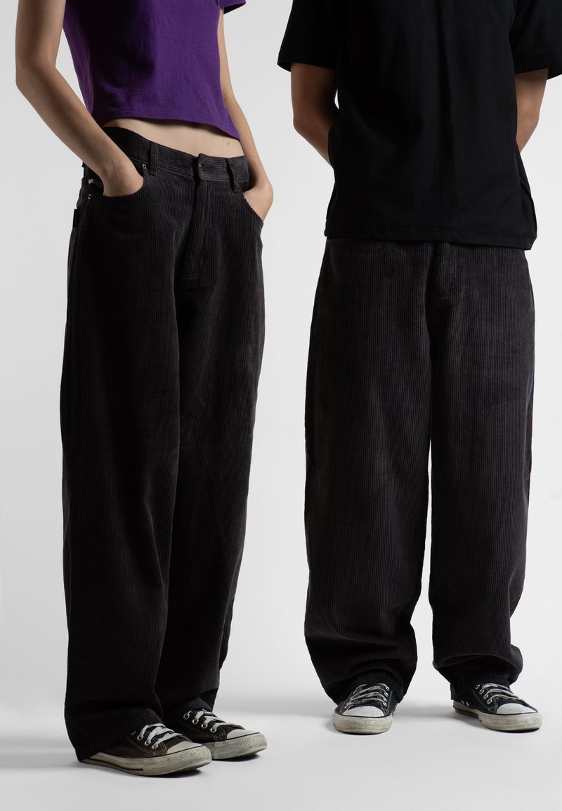 Pantaloni a gamba larga in corduroy neri con tasche anteriori, indossati da due persone. Uno indossa una maglietta corta viola, l'altro una maglietta nera.
