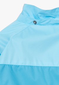 Světle modrá bunda s texturovaným povrchem, vyznačující se vysokým límcem, zapínáním na patentky a kontrastním barevným blokem na spodní části.