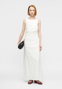 Λευκό μάξι φόρεμα από λινό με σχεδιασμό χωρίς μανίκια, εφαρμοστή σιλουέτα και διακριτική υφή. Συνδυασμένο με πλεκτή μαύρη τσάντα χειρός και καφέ φλατ παπούτσια.