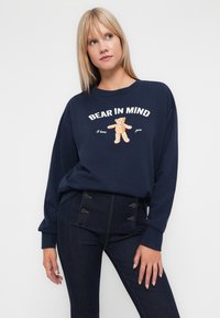 Navy sweatshirt with "BEAR IN MIND" text and a bear graphic. Long sleeves, crew neckline, and a relaxed fit. Worn with dark denim pants featuring button details.