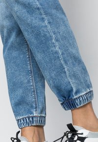 Dolní nohy mají na sobě světle modré džíny s elastickými manžetami a bílé tenisky s černými tkaničkami na jednoduchém světlém pozadí.