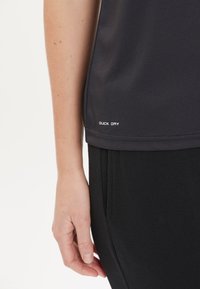 Svart snabbtorkande träningsskjorta med en slät textur. Har en nederkantsetikett med "QUICK DRY" i vitt. Kombinerad med svarta byxor, handen vilar.