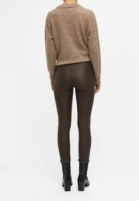 Nainen, jolla on päällään ruskeat istuvat nahkahousut, beige pitkähihainen neule ja mustat korotettu nilkkasaappaat, seisoo selkä kameralle valkoista taustaa vasten.