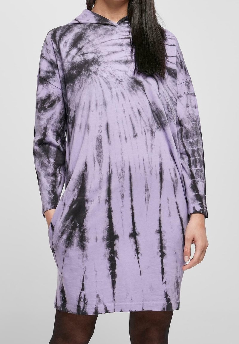Femme portant une robe à capuche tie-dye violet et noir à manches longues, debout les mains dans les poches sur un fond uni.