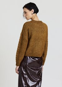 Brauner Pullover mit Zopfmuster, Rundhalsausschnitt und überschnittener Schulter, kombiniert mit einem glänzenden schwarzen Lederrock. Strukturierte Stoffdetails sichtbar.