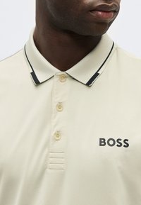 Κοντινό πλάνο ενός άντρα που φοράει μπεζ πόλο πουκάμισο με τρία κουμπιά, μαύρο περίγραμμα στο γιακά και το λογότυπο "BOSS" στο στήθος.