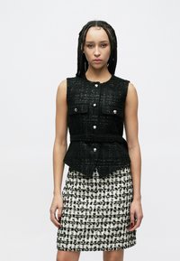 Schwarze Tweed-Weste mit ärmellosem Design, vorne mit Taschen und silbernen Knöpfen. Kombiniert mit einem schwarz-weiß karierten Rock.