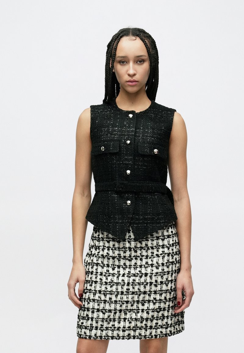 Schwarze Tweed-Weste mit ärmellosem Design, vorne mit Taschen und silbernen Knöpfen. Kombiniert mit einem schwarz-weiß karierten Rock.