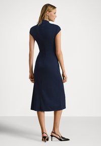 Navy oprijeta obleka s kratkimi rokavi, dolžine do kolen, gladke teksture, z visokim ovratnikom in zadrgo na hrbtni strani; kombinirana s črnimi salonarji z odprto peto.