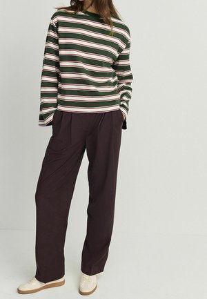 Personne portant un haut à manches longues rayé vert, blanc et rouge, un pantalon ample foncé et des baskets blanches, debout les mains dans les poches.