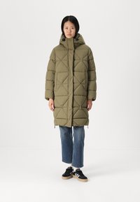 Olivenfarbener gepolsterter Mantel mit Kapuze, mit Rautensteppung und einem Frontreißverschluss. Getragen über blauen Jeans und schwarzen Sneakers.