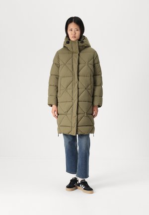 Olijfgroene gewatteerde jas met capuchon, voorzien van een ruitpatroon en een rits aan de voorkant. Draagt over blauwe jeans en zwarte sneakers.