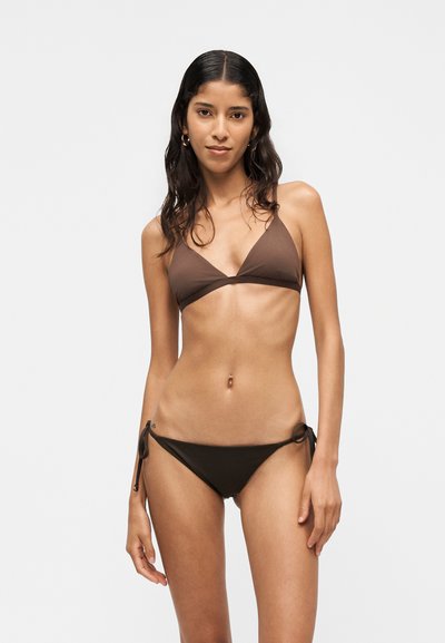 Jeune femme aux longs cheveux foncés portant un haut de bikini marron en forme de triangle et un bas de bikini assorti avec des liens sur les côtés, debout devant un fond clair uni.