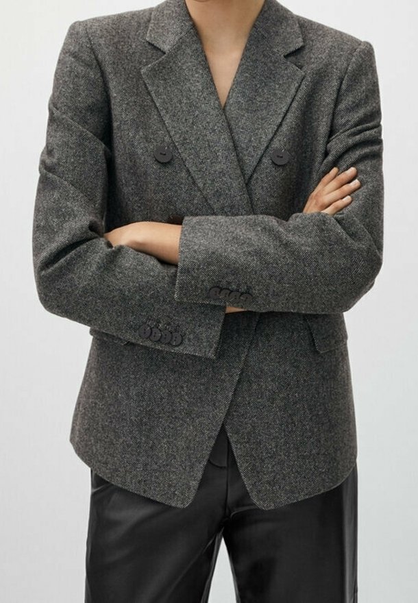 Personne portant un blazer gris croisé à boutons noirs, les bras croisés, associé à un pantalon en cuir noir sur un fond uni.