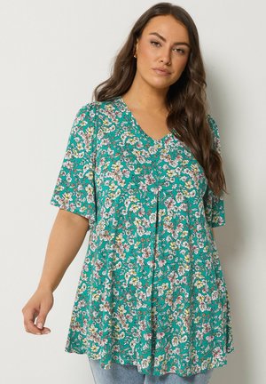 Kvinde med langt hår iført en løs, kortærmet teal tunika med flerfarvet blomsterprint og V-hals, stående mod en ensfarvet væg.