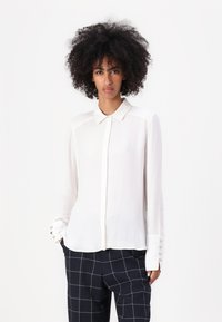 Blusa blanca de botones de manga larga en una tela ligera, con cuello y botones decorativos en los puños, combinada con pantalones de cuadros en azul marino.