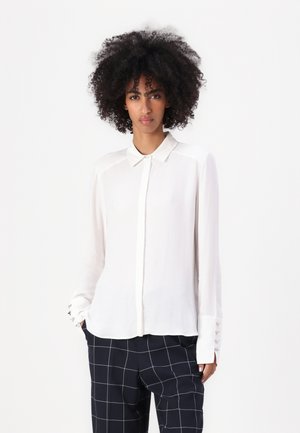 Blusa bianca a maniche lunghe con bottoni in un tessuto leggero, con colletto e bottoni decorativi ai polsini, abbinata a pantaloni in plaid navy.